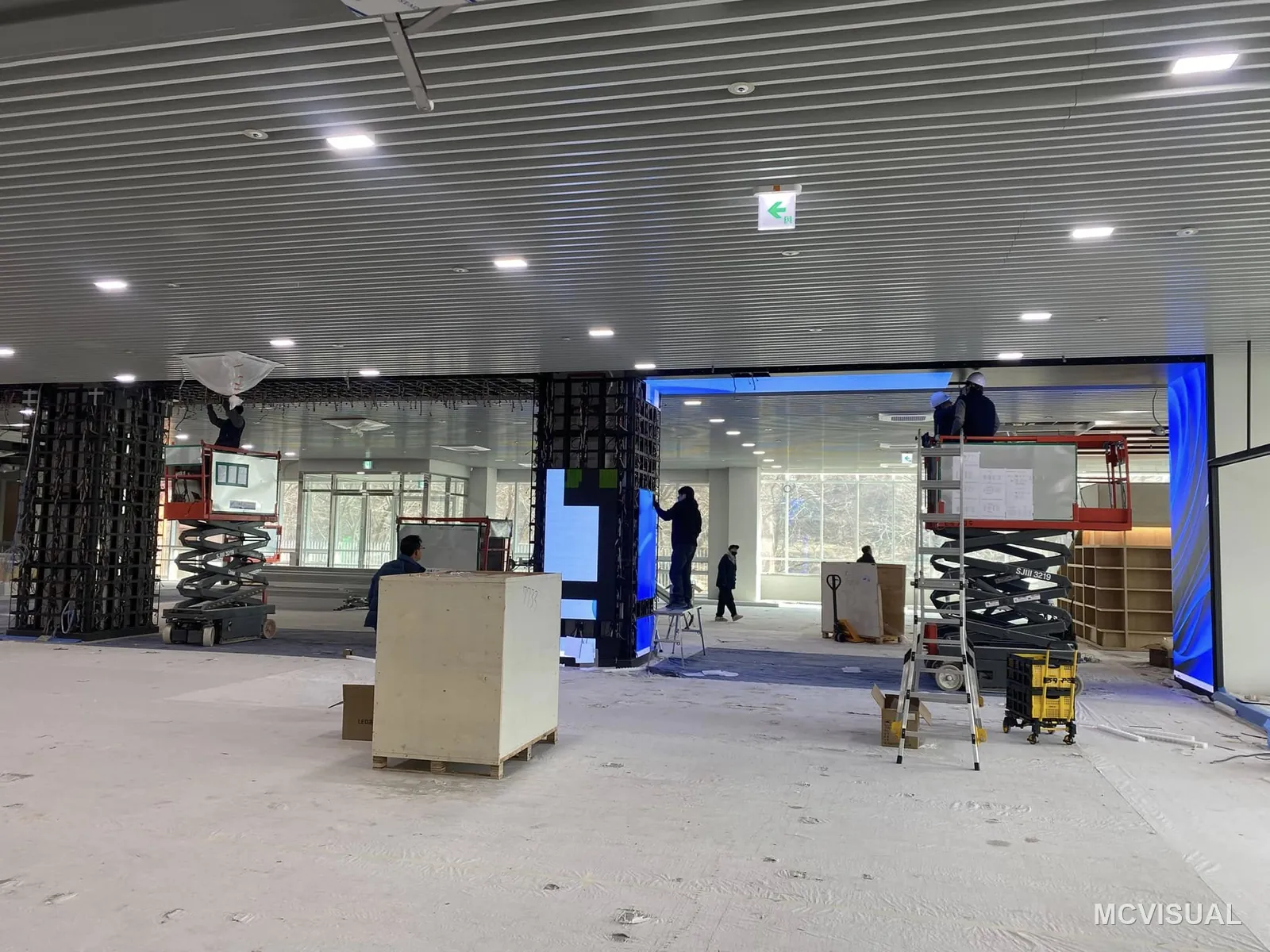 Retail concourse, South Korea — The concourse mid-fit-out, screens going up from scissor lifts while other trades work around them. Finishing inside the